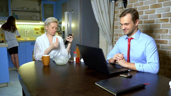 Um homem que trabalha em casa no laptop na cozinha, esposa come — Fotografia de Stock