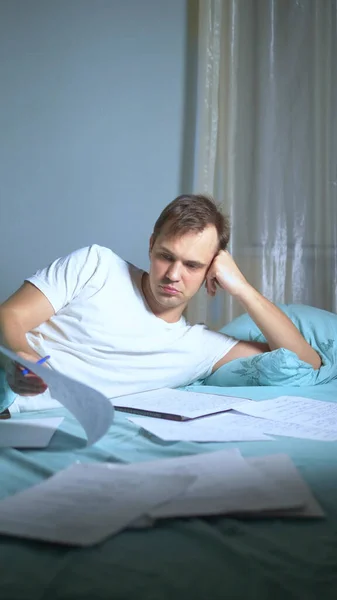 Vertikalt skott. En man skriver på pappershandlingar liggande i sängen. arbete i hemmet. — Stockfoto