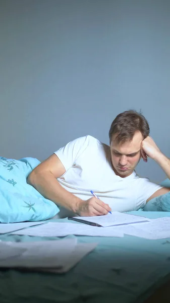 Plan vertical. Un homme écrit sur des papiers couchés au lit. travail à domicile . — Photo