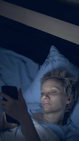 Вертикально. женщина, страдающая бессонницей, пользуется смартфоном, когда лежит в постели — стоковое фото