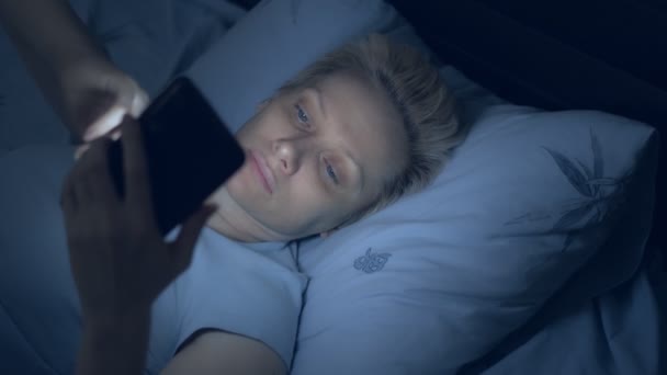 O femeie care suferă de insomnie folosește un smartphone în timp ce stă în pat în întuneric — Videoclip de stoc