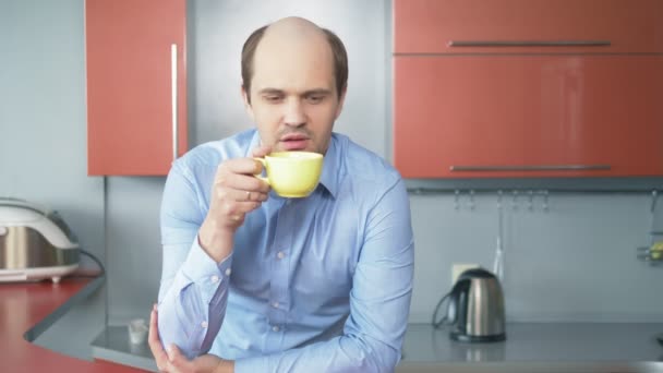 Łysiejący młodzieniec pijący herbatę w kuchni. patrząc w kamerę — Wideo stockowe