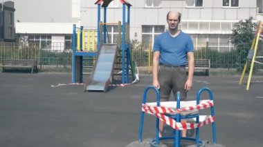 Garip, kel, çocuksu bir adam boş bir oyun parkında duruyor.