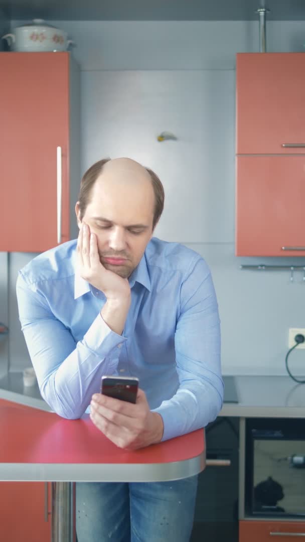 Pionowo. łysiejący młody człowiek używa smartfona w kuchni w domu. — Wideo stockowe