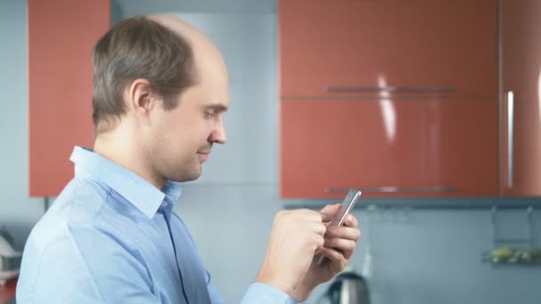 Ο φαλακρός νεαρός χρησιμοποιεί ένα smartphone στην κουζίνα στο σπίτι. — Αρχείο Βίντεο