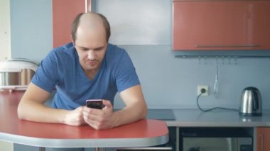 Kel genç adam evdeki mutfakta akıllı telefon kullanıyor..