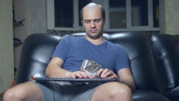 Καραφλός άνθρωπος δακτυλογραφεί σε ένα πληκτρολόγιο, κάθεται στο σπίτι σε μια πολυθρόνα και τρώει μάρκες — Αρχείο Βίντεο