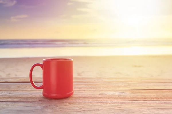 Red coffee cup on wood table at sunset or sunrise beach — Stock Photo, Image
