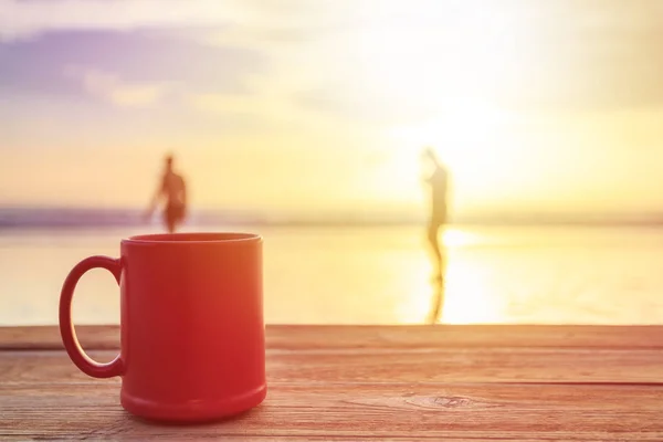 Red coffee cup on wood table at sunset or sunrise beach — Stock Photo, Image