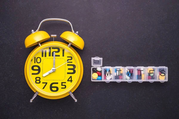 7 gün plastik hap organizer ve sarı alarm clock karanlık tabl üzerinde — Stok fotoğraf