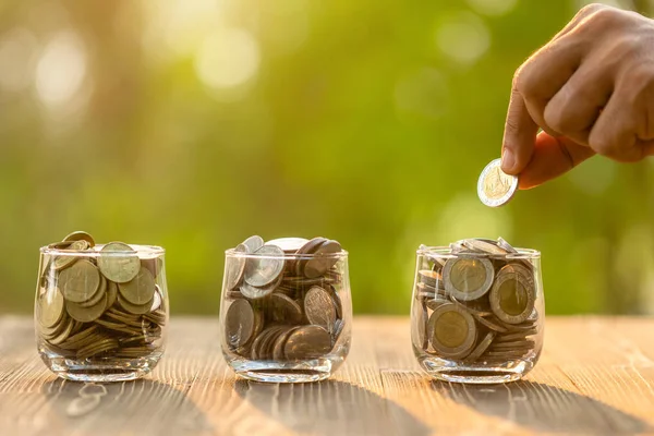 Hand putting coins into clear money jar on wooden table with gre — Stock Photo, Image