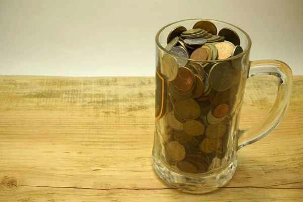 Glass mug with different metal coins on a wooden table on the right and left-background for the inscription
