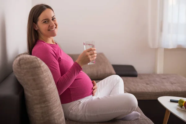 美しいです妊娠中の女性楽しんでいます飲料水とともに彼女の家でソファに座って — ストック写真