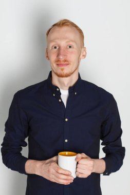 Bir fincan çay veya kahve ile genç bir adam. O memnun. Beyaz arka plan. Kızıl saçlı erkek beyaz kupa ile.