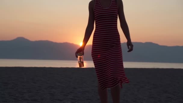 Menina sexy magro em vestido listrado vermelho andando lentamente ao longo da praia com um copo de mojito fresco, silhueta de menina bonita no incrível rosa e laranja fundo do pôr do sol. Férias de verão, festa na praia — Vídeo de Stock