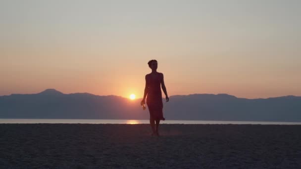 Uzun yaz elbisesi içinde, elinde taze kokteyl ya da mojito tutan ve deniz günbatımında kameraya poz veren çekici çıplak ayaklı bir kız. Genç bir kızın unutulmaz yaz maceraları — Stok video