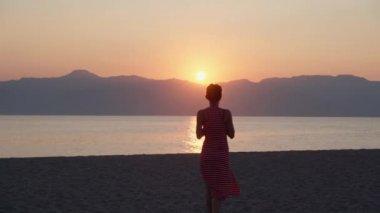 Sıska kızın uzun kırmızı yaz elbisesiyle deniz suyuna yaklaşıp güzel, doğal günbatımının tadını çıkardığını görüyorum. Deniz sahilinde rahatlamak, yürümek ve taze içecekler içmek, plaj partisi konsepti.