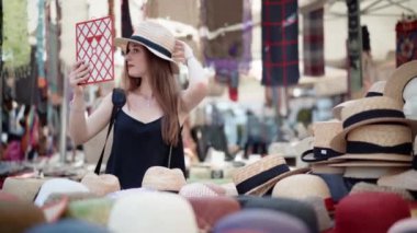 Genç bir kadın bit pazarında panama şapkası deniyor ve aynaya bakıyor. Yaz moda trendleri, piyasa ticareti ve güzellik konsepti 