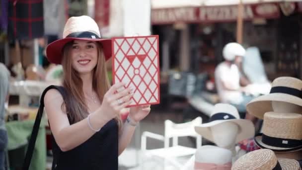 Attractive tourist girl trying on colorful summer hat at the local craft market, looking into the mirror and smiling on camera. Protective head ware for summer vacation, beauty and fashion — Stock Video