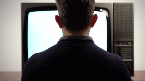 Вид сзади на мужчину, сидящего перед винтажным экраном телевизора с помехами от шума и смотрящего трансляцию. Медиапередачи на ретро-ТВ 90-х годов. Старая телевизионная концепция — стоковое видео