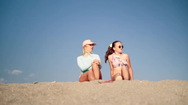 Ciepły słoneczny dzień na plaży surfingowej, atrakcyjna młoda blondynka w kapeluszu ochronnym, siedząca na piasku ze swoją nastoletnią córką w okularach i bikini w kwiatowym wzorze. Szczęśliwa rodzina ma przyjemne — Wideo stockowe