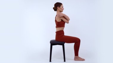 Spor giyimli güzel bir kız izole edilmiş beyaz arka planda siyah bir taburede oturuyor ve omurgasını güçlendirmek için egzersiz yapıyor. Fitness kızı elleriyle omuzlarına sarılıyor ve eğiliyor.