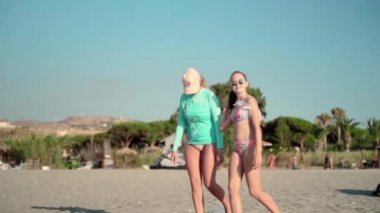 Deniz sahilinde aile tatilleri, kızıyla birlikte deniz kıyısında ağır çekimde yürüyen anne, gülümseyen ve eğlenen öğleden sonraları. Koruyucu gözlüklü kız güneş banyosu yapıyor, anne şapkalı ve uzun kollu.