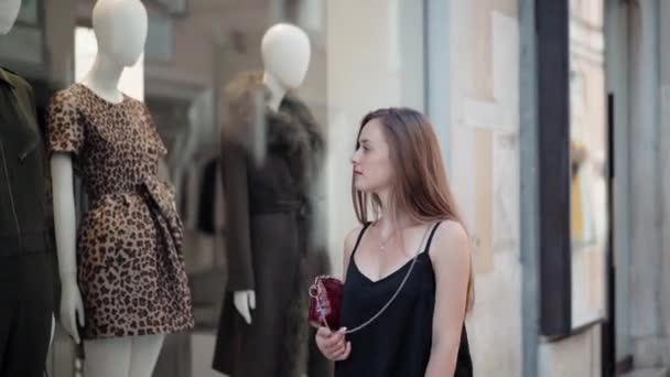 Jolie fille aux longs cheveux roux marchant dans la rue et regardant les vitrines éclairées avec une belle collection de vêtements de luxe. Grandes ventes dans le magasin, femme accro au shopping regardant avec plaisir — Video