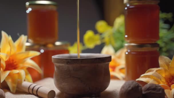 Miel frais délicieux goutte à goutte dans un bol en bois, produit naturel doré avec des vitamines. Produit apicole pur coulant sur fond floral étonnant avec trempette en bois et bocaux de miel — Video