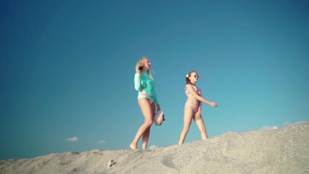 驚くべき青い空の背景と幸せな家族はゆっくりとした動きで海のビーチに沿って裸足で歩き、男性のカイトサーファーで通過します。リラックスした散歩ブロンド母と若いです可愛いです娘,夏 — ストック動画