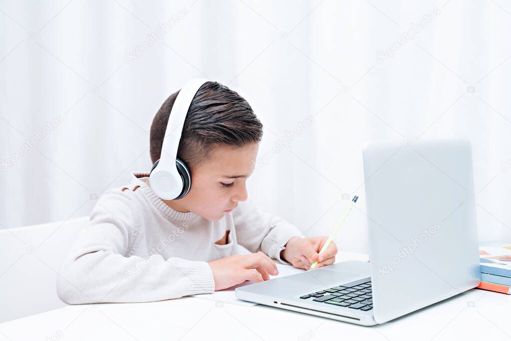  Child study online sitting on the a computer with headphones. The child writes in a notebook