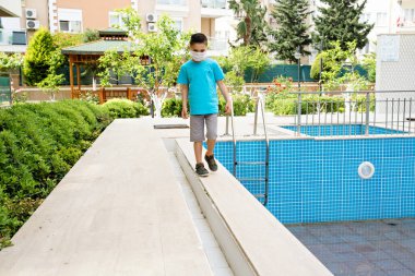 Tıbbi maskeli bir çocuk boş bir yüzme havuzunun yanından geçiyor. 2020 yazını düşün. Turizm yok. 