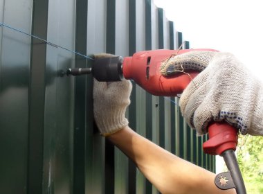 Metal bir çit yerleştirilmiş. Eldiven giymiş, kırmızı matkap kullanan bir işçi bahçesinde yeni bir metal, kıvrımlı çit, yeşil bir çit inşa ediyor. Yakın plan fotoğraf..