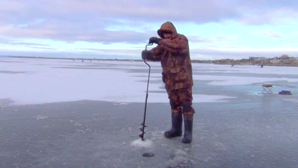 A fisherman in winter in warm orange, spotted clothes, on a frozen river drills a fishing hole with a device on the winter ice — Stock Footage Fisherman Winter Warm Orange Spotted Clothes Frozen River Drills Fishing Royalty Free Stock Video