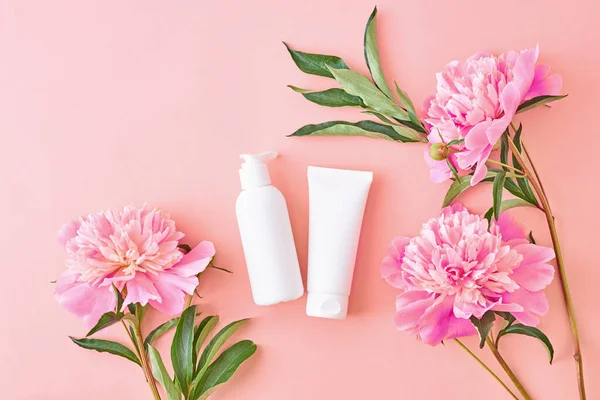 Flat lay composition with cosmetic products, mockup white tubes and pink peonies on a pink background