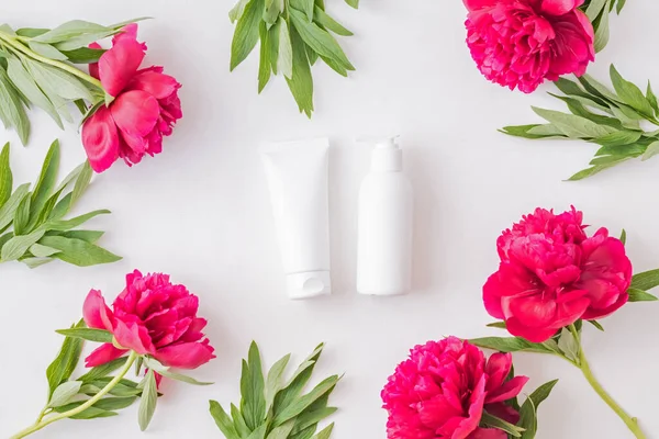 Flat lay composition with cosmetic products, mockup white tubes and red peonies on a white background