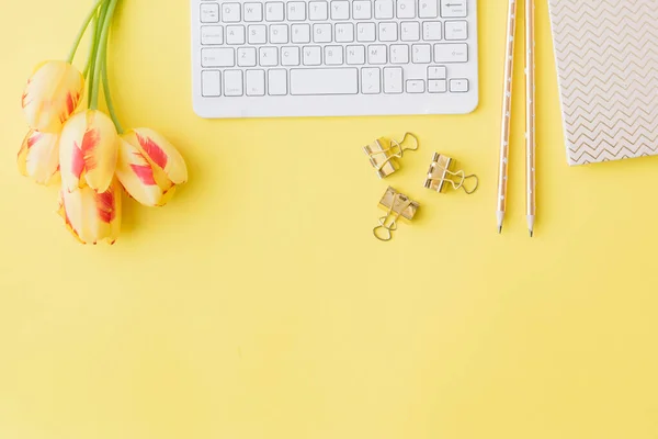Flat lay blogger or freelancer workspace with a notebook, keyboard and yellow tulips on a yellow background
