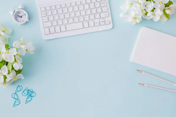 Flat lay blogger or freelancer workspace with a notebook, keyboard and white spring flowers on a blue background