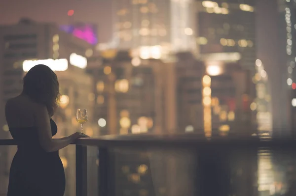 Bella giovane donna sul balcone in un abito nero con un bicchiere di vino sullo sfondo di una città notte — Foto Stock