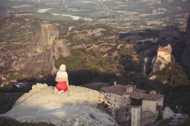 Yalnız kız bir kırmızı elbise ve kaya kenarında eşarp ve Meteora manastırları dua ediyor. Kadın rock ve Yunanistan Thessaly'deki/daki oteller Meteora manastırları