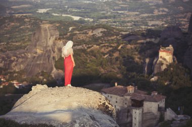 Yalnız kız bir kırmızı elbise ve kaya kenarında eşarp ve Meteora manastırları dua ediyor. Kadın rock ve Yunanistan Thessaly'deki/daki oteller Meteora manastırları