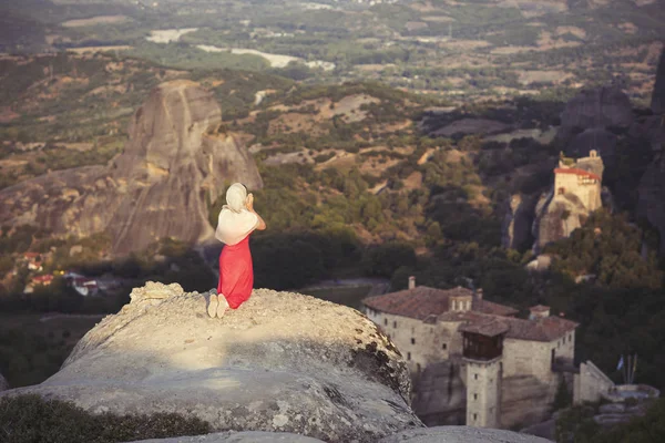 Yalnız kız bir kırmızı elbise ve kaya kenarında eşarp ve Meteora manastırları dua ediyor. Kadın rock ve Yunanistan Thessaly'deki/daki oteller Meteora manastırları