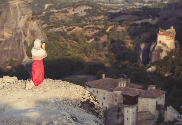 Menina sozinha em um vestido vermelho e cachecol na borda da rocha e reza nos mosteiros de Meteora. Feminino na rocha e mosteiros de Meteora, na Grécia, na Tessália — Fotografia de Stock