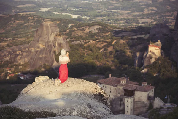 Egyedül lány egy piros ruha és kendő a szikla szélén, és imádkozik a Meteórák kolostoraihoz. Nő a rock és a Meteórák kolostoraihoz Thesszália, Görögország — Stock Fotó