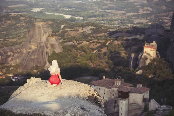 Yalnız kız bir kırmızı elbise ve kaya kenarında eşarp ve Meteora manastırları dua ediyor. Kadın rock ve Yunanistan Thessaly'deki/daki oteller Meteora manastırları