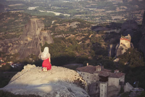 Yalnız kız bir kırmızı elbise ve kaya kenarında eşarp ve Meteora manastırları dua ediyor. Kadın rock ve Yunanistan Thessaly'deki/daki oteller Meteora manastırları