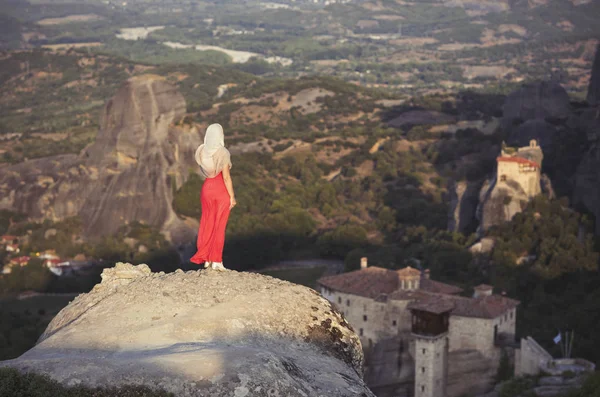 Yalnız kız bir kırmızı elbise ve kaya kenarında eşarp ve Meteora manastırları dua ediyor. Kadın rock ve Yunanistan Thessaly'deki/daki oteller Meteora manastırları