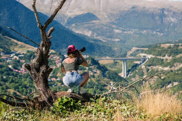 Jovem fotógrafa feminina com câmera profissional perto de grande árvore velha em frente à aldeia grega. Uma menina com câmera em frente à aldeia de Metsovo, na Grécia — Fotografia de Stock