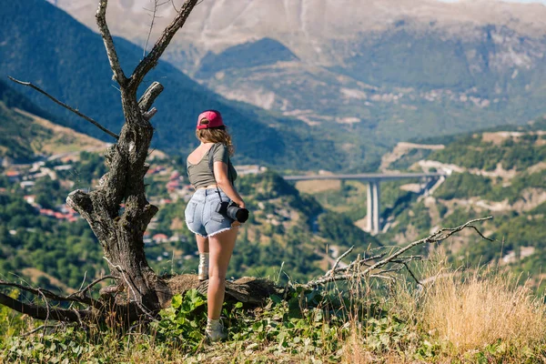 Jovem fotógrafa feminina com câmera profissional perto de grande árvore velha em frente à aldeia grega. Uma menina com câmera em frente à aldeia de Metsovo, na Grécia — Fotografia de Stock