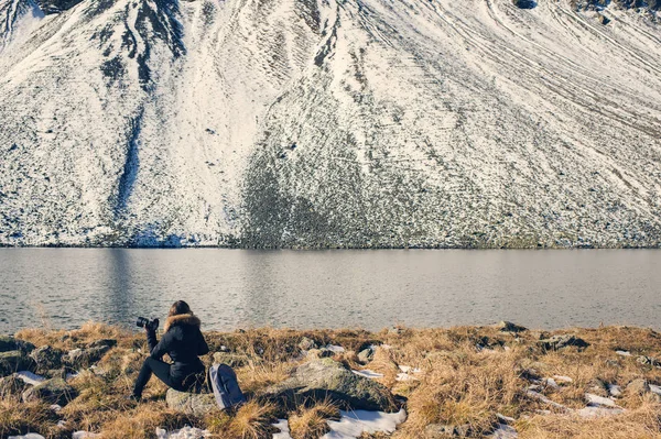 Uma fotógrafa com câmera em uma jaqueta de inverno com pêlo fica em frente às montanhas de neve e um lago na Suíça. Fluela passa na Suíça no inverno . — Fotografia de Stock